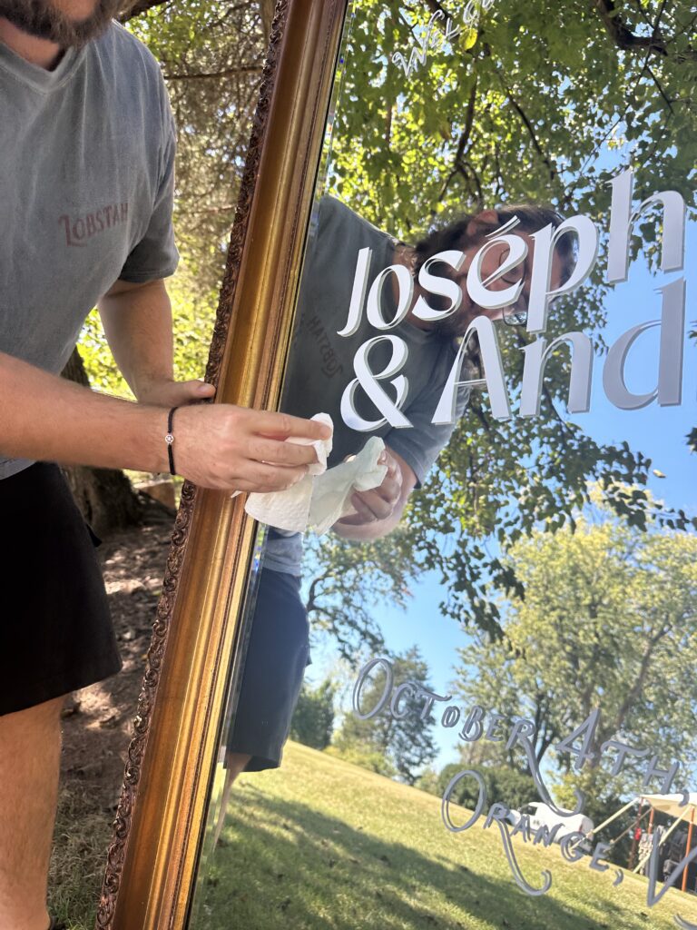 A person polishing a gold-framed mirror lettered in white vinyl with a couple's names, outdoors under a tree