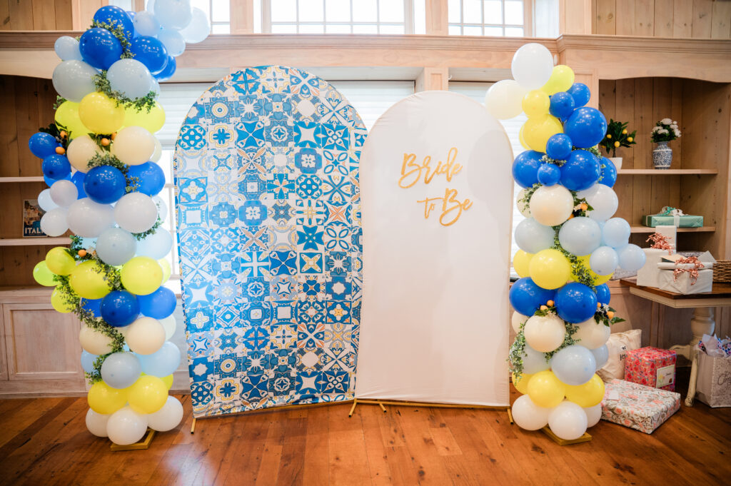 Two freestanding arch backdrop panels, one in blue-and-white tile print and one lettered in gold, with a full-height balloon column in blue, yellow, white and cream either side, threaded with greenery and faux lemons