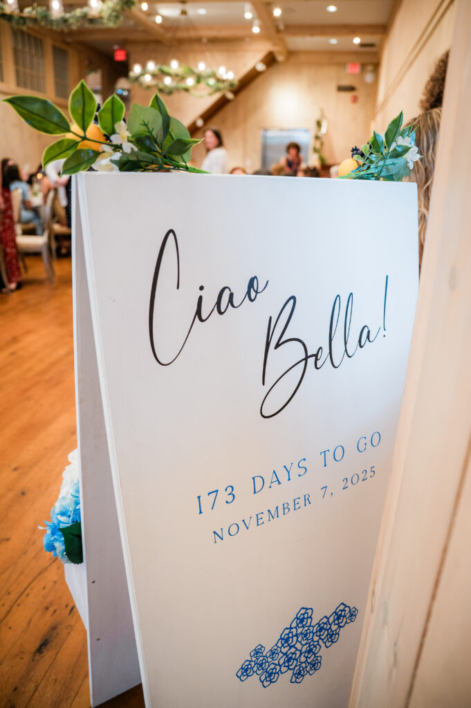 A white A-frame sign lettered in black script with a countdown, topped with greenery, faux lemons and white blossom