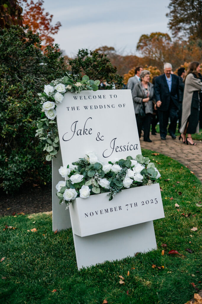 A white freestanding welcome sign on grass at an outdoor wedding, its flower box filled with white roses and eucalyptus