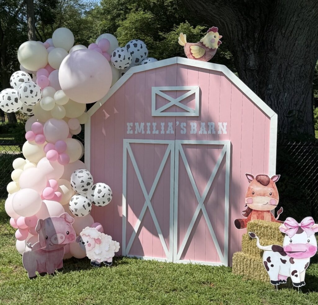 A pink barn photo backdrop on a lawn, lettered EMILIA'S BARN across the front, with a balloon garland in pink, cream and cow print and a chicken cutout perched on the roof