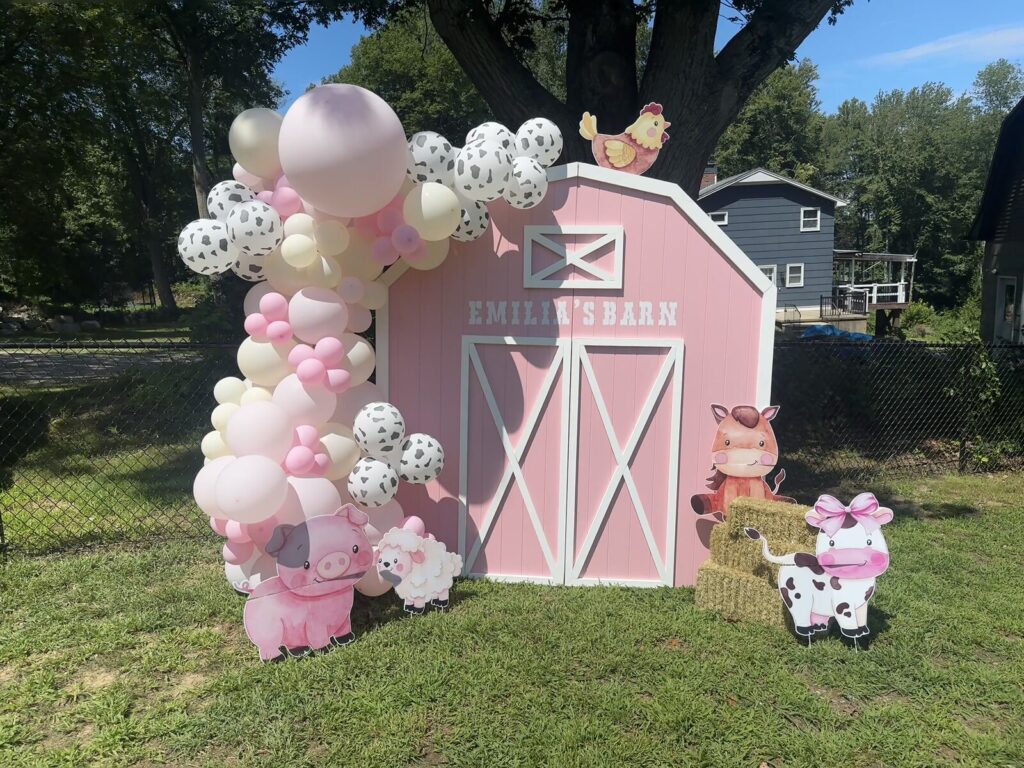 The full barnyard setup on a lawn under a tree — pink barn backdrop, balloon garland, hay bale and cutouts of a pig, a sheep, a horse and a cow