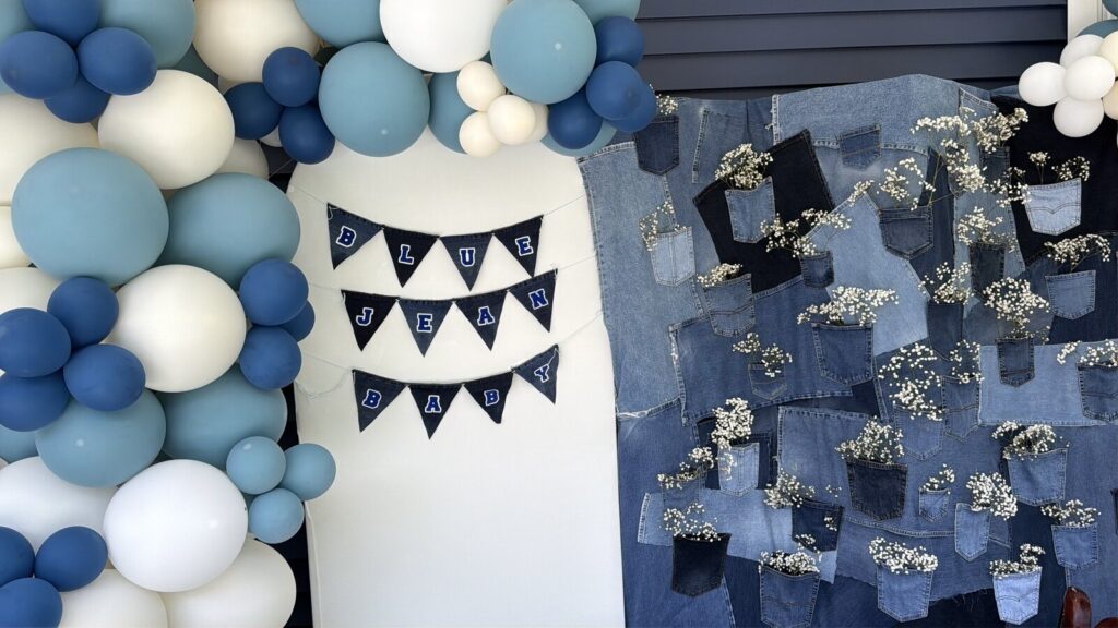 A denim patchwork backdrop with baby's breath tucked into the jean pockets, a white arch with navy BLUE JEAN BABY bunting, and a balloon garland in blue, dusty blue and cream