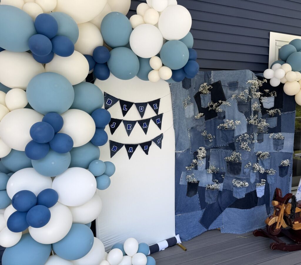 The full Blue Jean Baby setup on a deck — denim pocket backdrop, white arch with navy bunting, blue and cream balloon garland and a wooden rocking horse