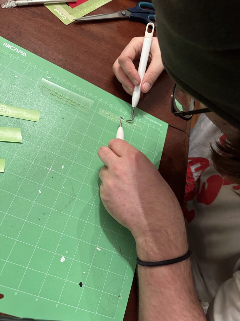 Two hands weeding vinyl lettering off a green cutting mat with a fine hook tool, offcuts and craft scissors around them