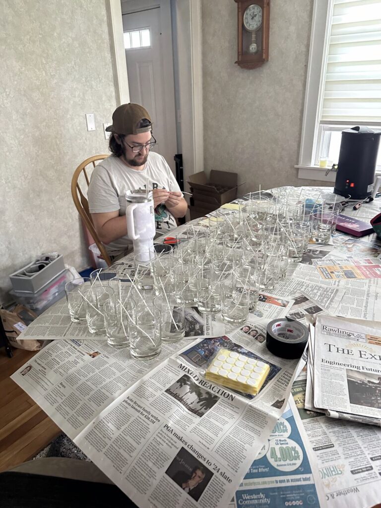 A hand setting a wick into a glass vessel over a newspaper-covered table