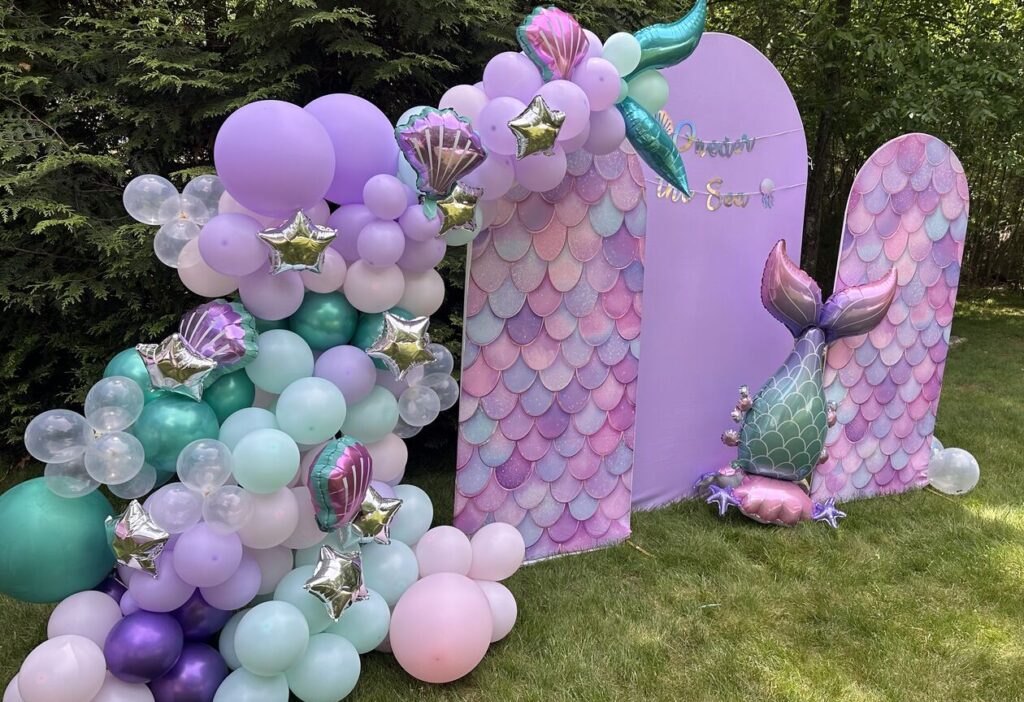 A mermaid-themed backdrop with a pastel balloon garland on a lawn