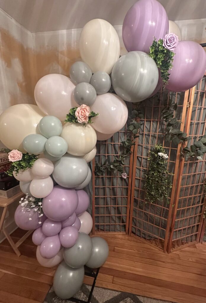 A balloon garland in lilac, cream, grey and white with pink flowers and greenery tucked through it, running up a lattice panel
