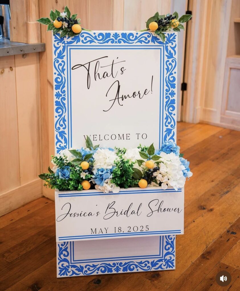 A white welcome sign with a blue tile-pattern border and a built-in flower box of blue and white hydrangea, greenery and faux lemons
