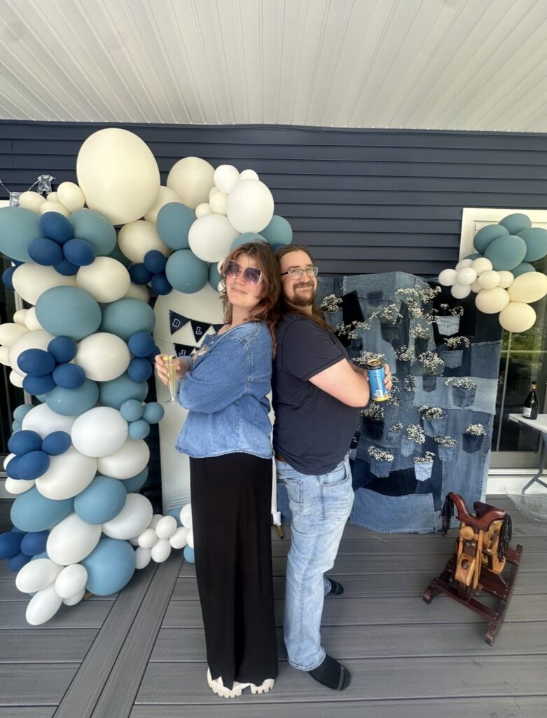 Vicky and Dylan standing back to back with their arms folded, mock-serious, in front of the balloon garland and denim backdrop they built for a Blue Jean Baby shower