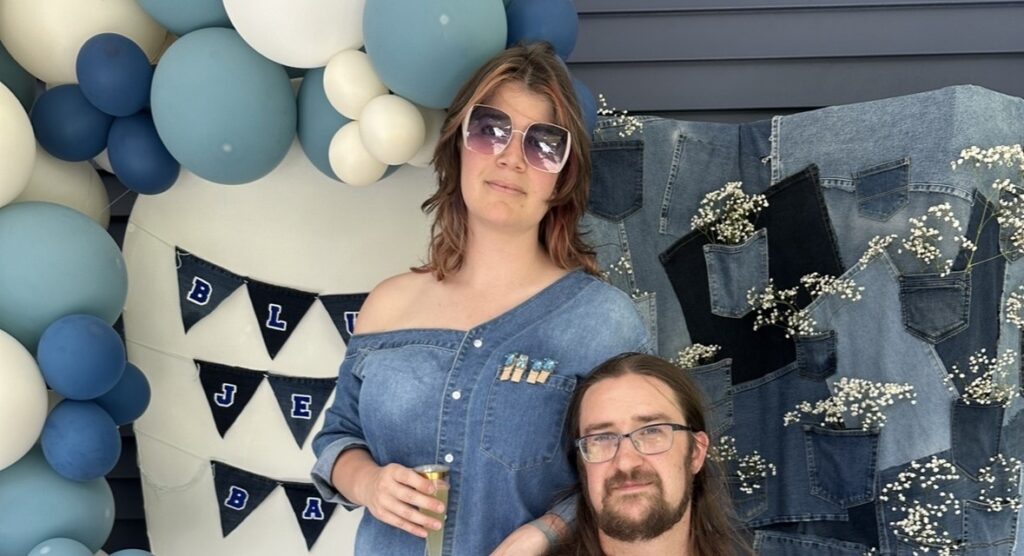 Vicky and Dylan in front of a baby shower setup they built — a blue and cream balloon garland, a denim patchwork backdrop with baby's breath tucked into the pockets, and a Blue Jean Baby banner