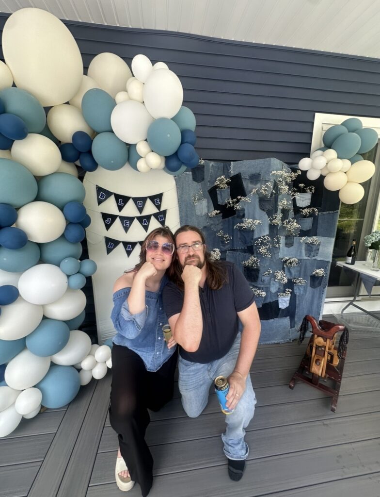 Vicky and Dylan crouching together, chins on their fists, in front of a finished baby shower setup — a blue and cream balloon garland, a denim patchwork backdrop with baby's breath tucked into the pockets, and a Blue Jean Baby banner