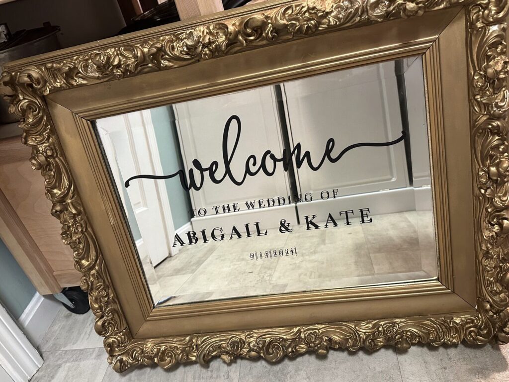 A welcome sign for a wedding, hand lettered in black script directly onto a mirror in an ornate carved gold frame