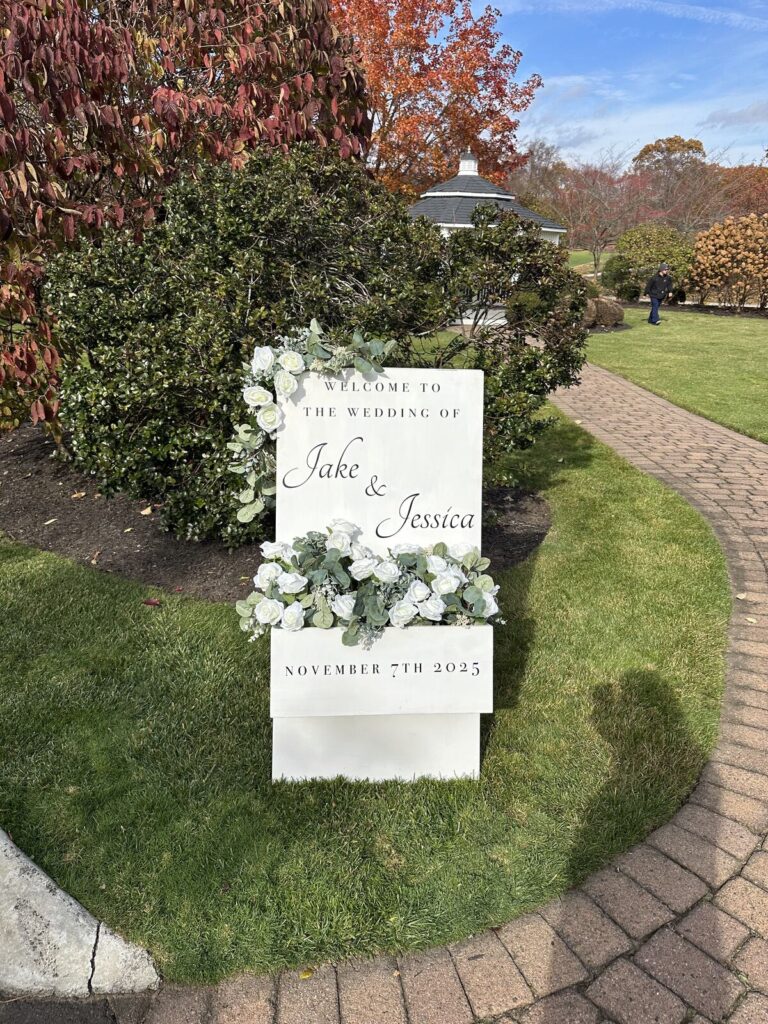 A tall wedding welcome sign in a garden, lettered in script and dressed with white florals