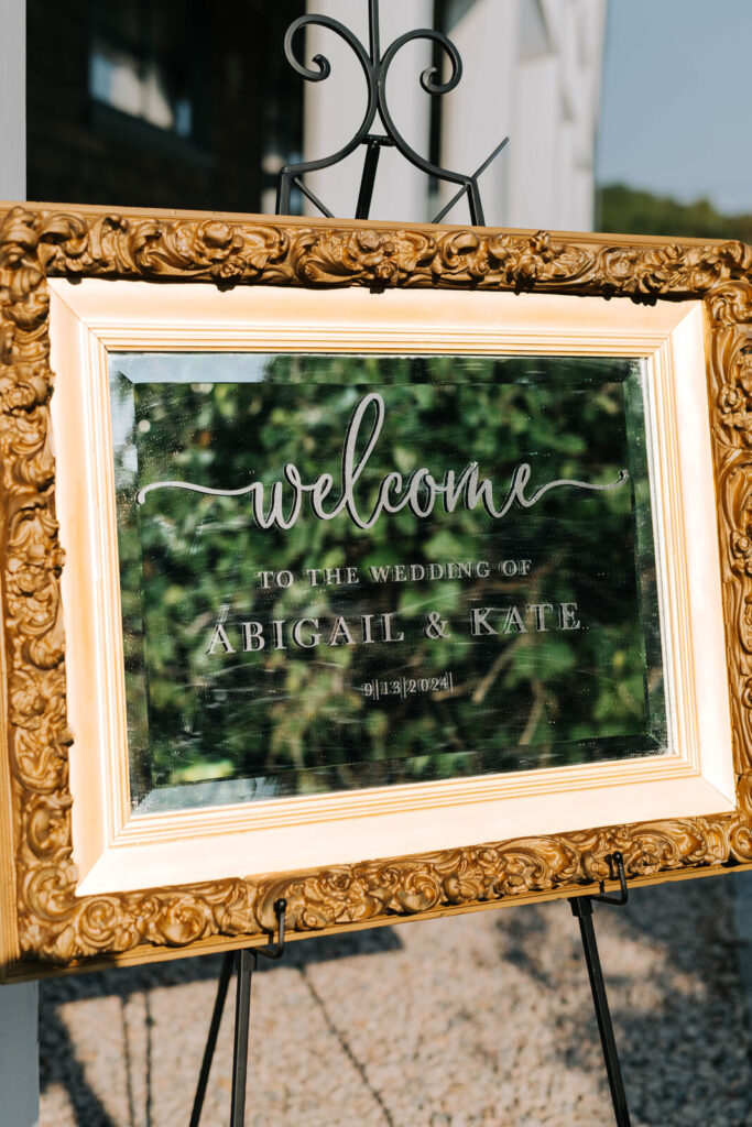 A gold-framed mirror welcome sign for a wedding, standing outdoors on an easel